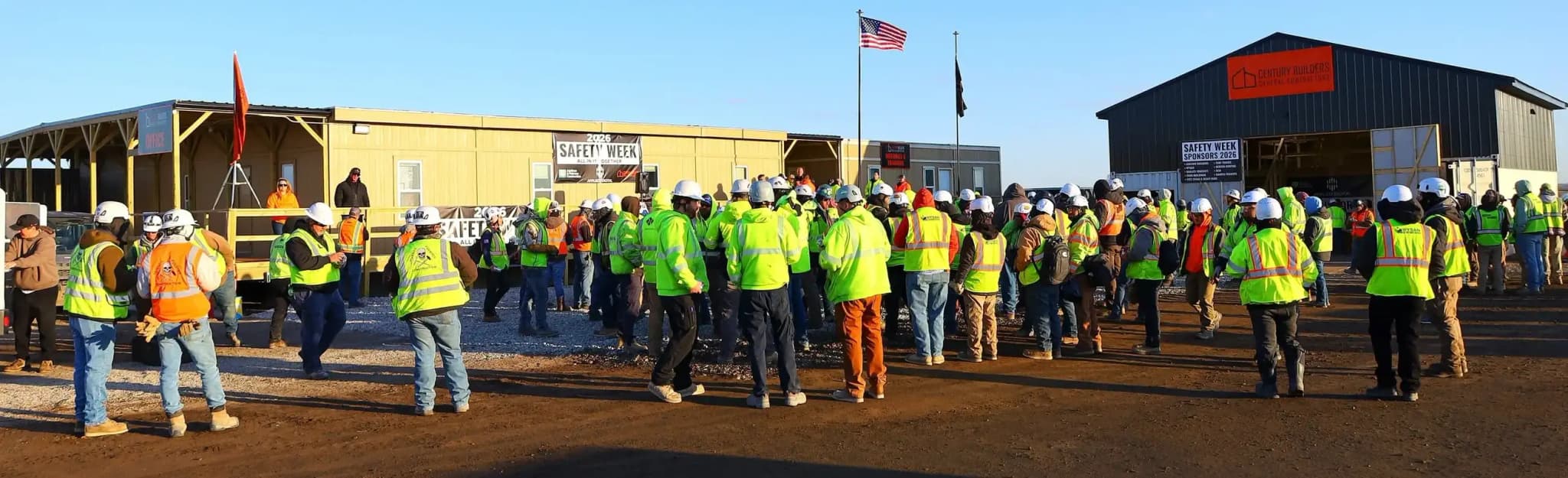 Century Builders crew and trade partners gathered outdoors for the 2026 Safety Week kickoff at the Harwood data center site, with the American flag flying and jobsite buildings behind them