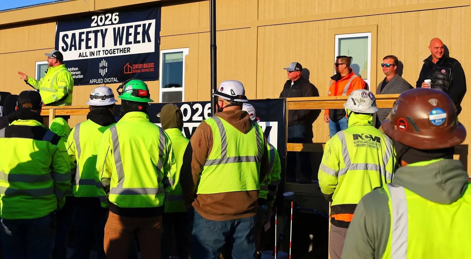 A speaker addresses the crew beneath the 2026 Safety Week banner, which carries the Century Builders logo, at the Harwood data center site